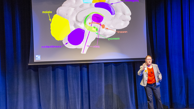 Dr. Gillian Beauchamp stands on stage in front of a projector slide identifying various areas of the brain triggered by addiction.